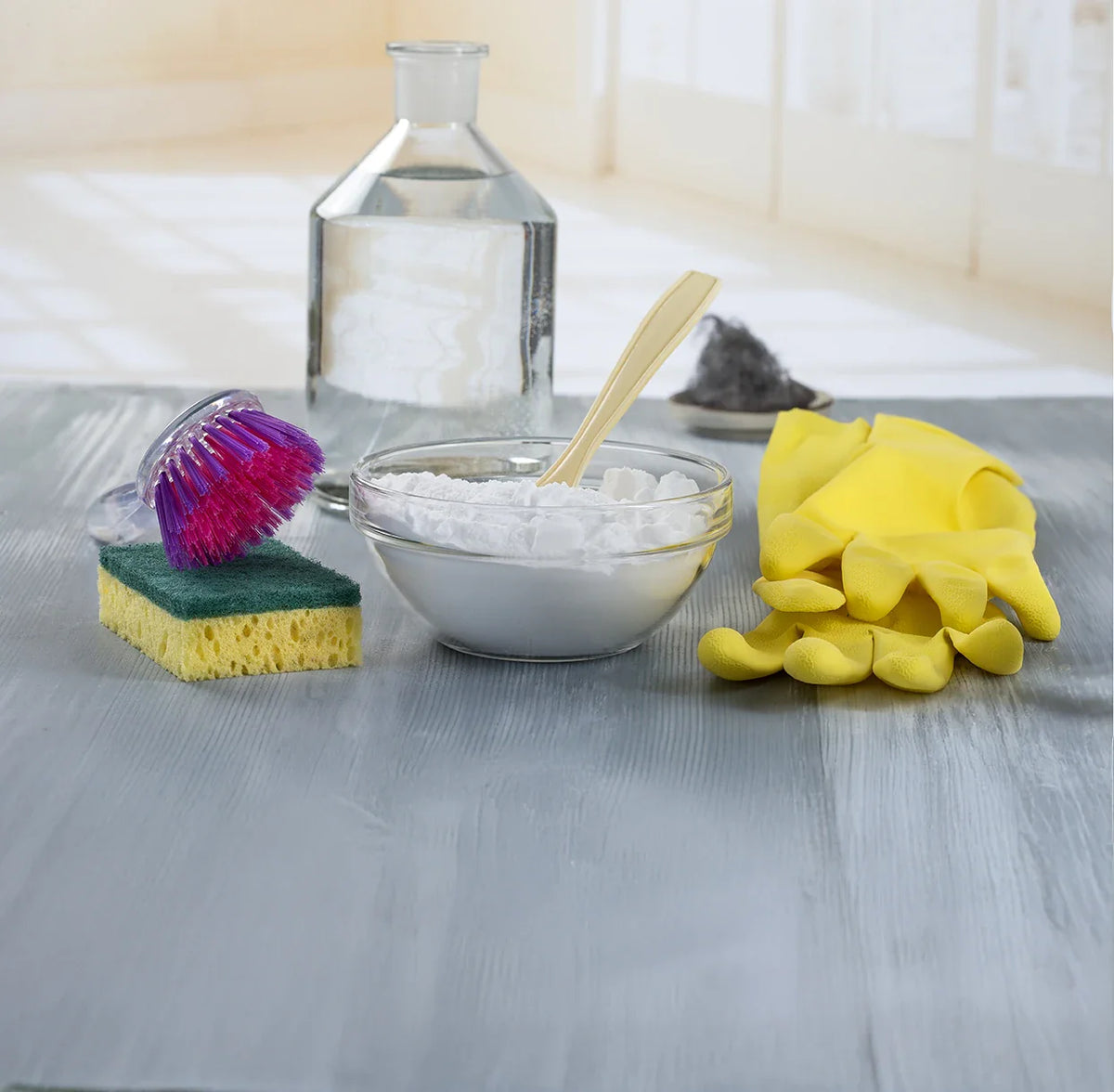 Reinigungsutensilien auf einem Tisch: Eine Schüssel mit Natron und einem Löffel, eine Flasche Essig, ein gelber Reinigungshandschuh, ein Schwamm mit einer Bürste mit pinken Borsten sowie ein kleiner Haufen Stahlwolle im Hintergrund.