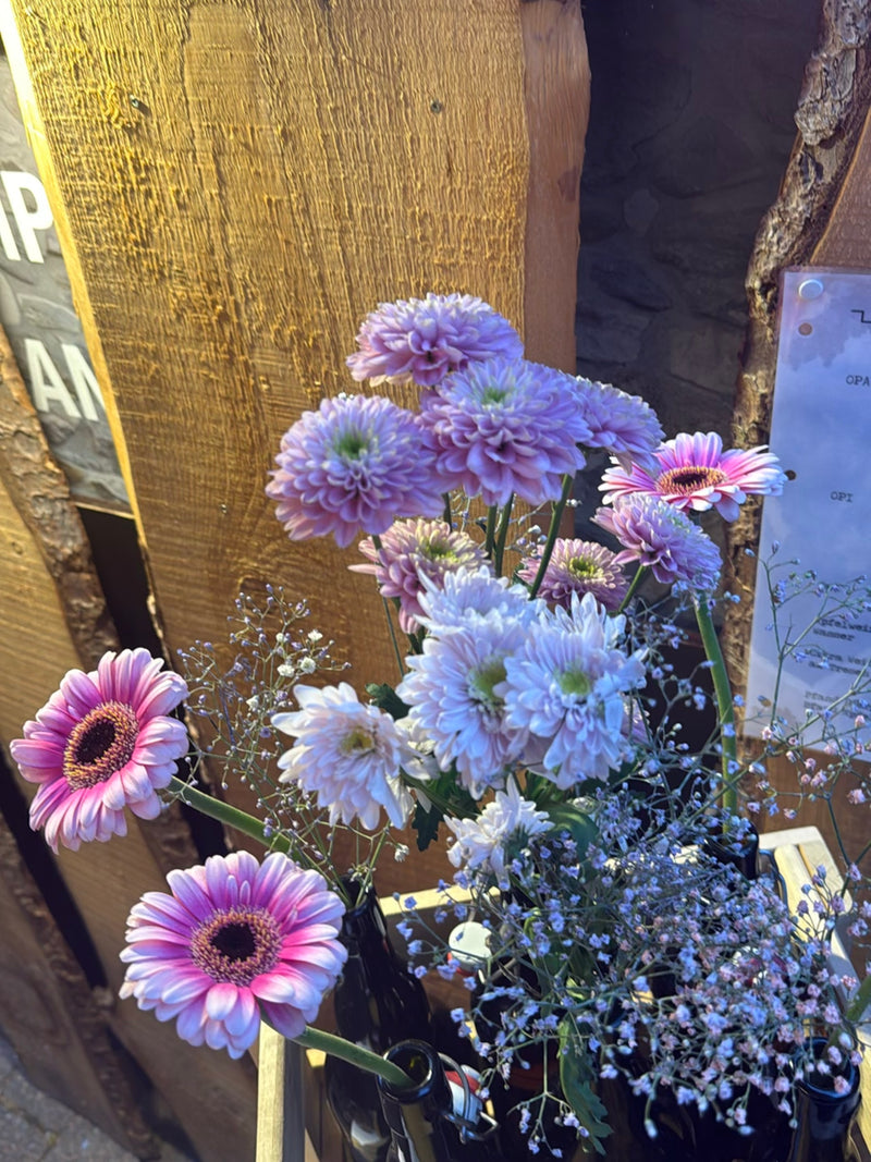 Plusieurs fleurs de gerbera roses se tiennent avec d'autres fleurs dans des bouteilles devant un mur en bois.