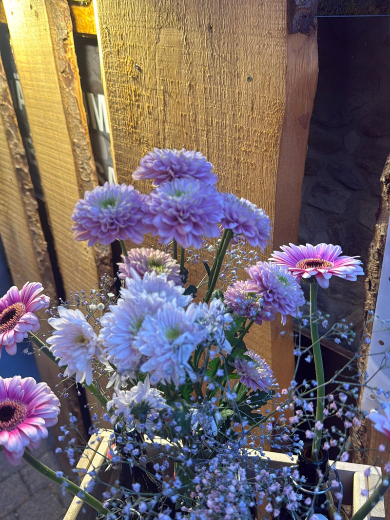 On voit un bouquet de fleurs coloré avec des gerberas et d'autres fleurs devant un mur en bois. Les plantes sont dans des récipients.